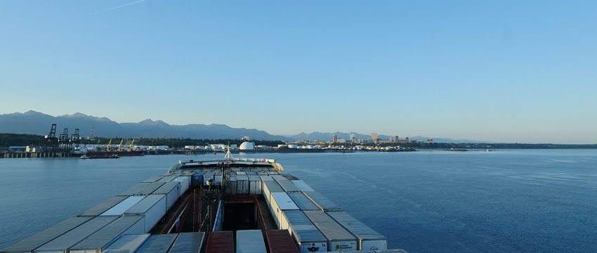 View of a cargo ship docked at a port with containers and a city skyline in the background.