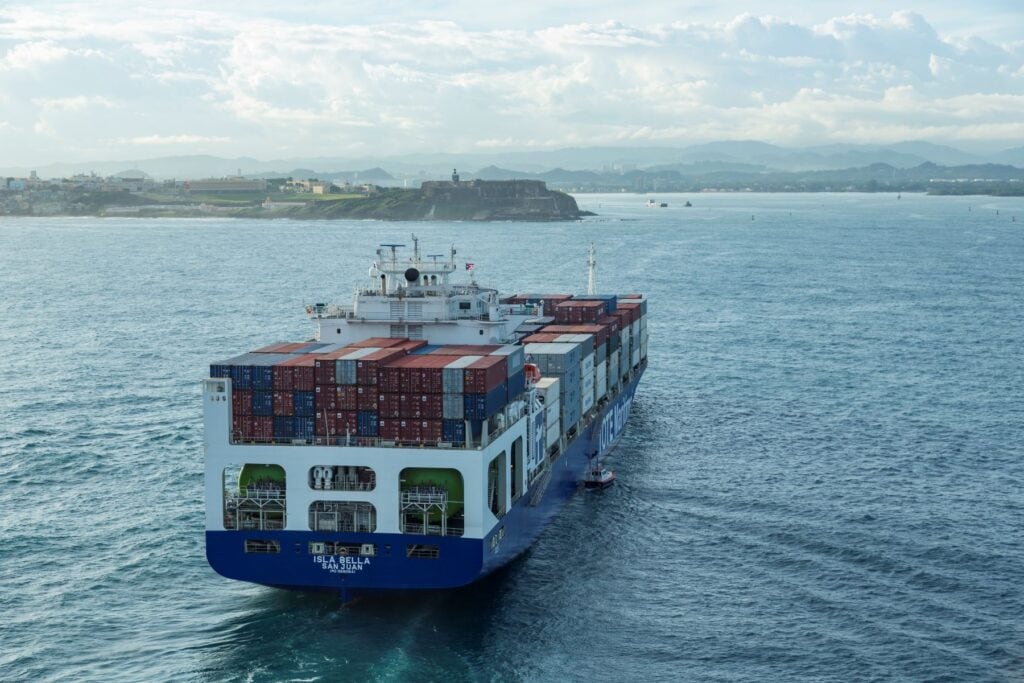 Container ship sailing on the water with a cityscape in the background.