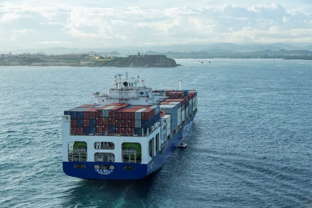 Container ship sailing on the water with a cityscape in the background.