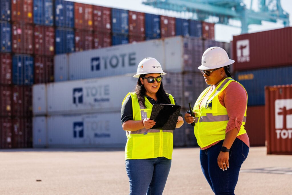 Workers in safety vests and helmets discussing at a shipping yard with stacked containers in the bac.