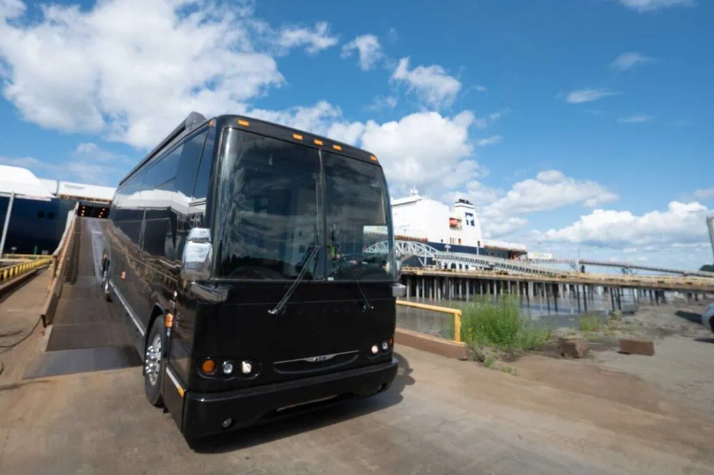 Black tour bus parked near a marina with yachts and boats in the background under a partly cloudy sk.