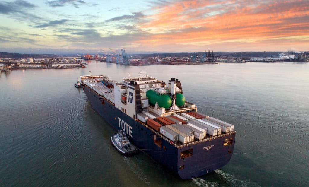 Tote cargo ship with containers and green tanks sailing in a harbor at sunset.