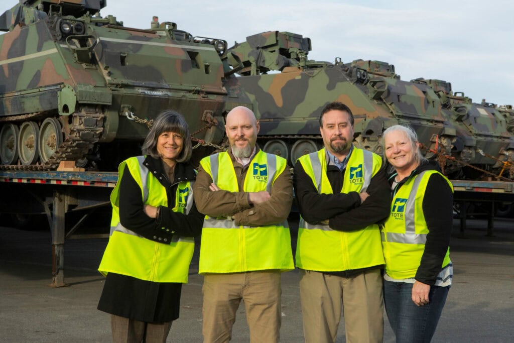 Military personnel in high-visibility vests standing in front of tanks on a flatbed trailer.