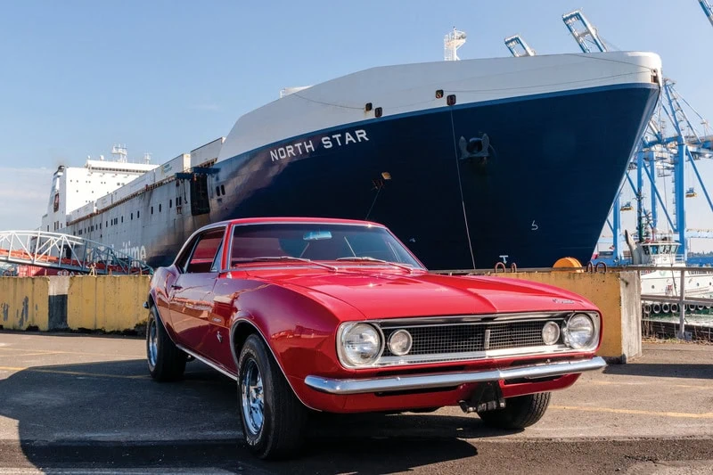 Vintage red muscle car parked near a large cargo ship at a port.