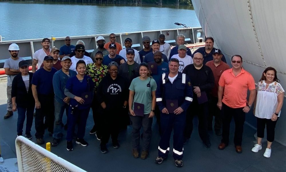 A boat crew gathered on the deck with a large ship in the background.