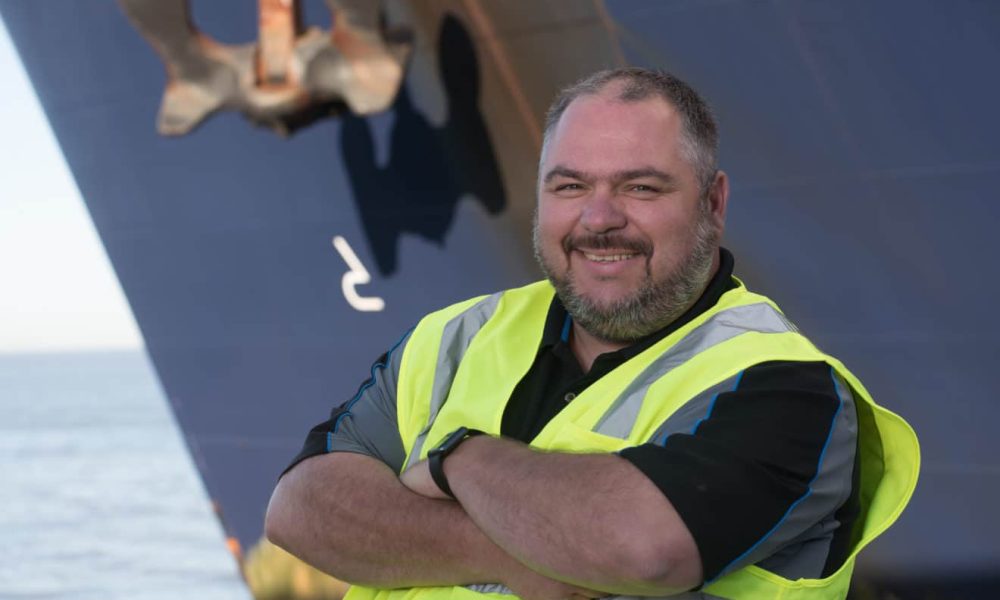 Man in high-visibility vest smiling with ship in background.