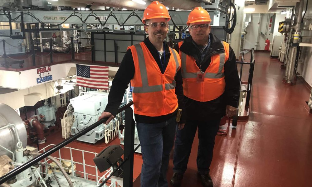 Two men in orange safety vests and helmets on an industrial balcony.