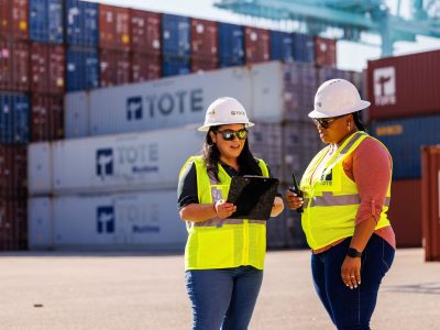 Ketterman_250729_3326-web – TOTE Group Workers in safety vests and helmets discussing at a shipping yard with stacked containers in the bac.