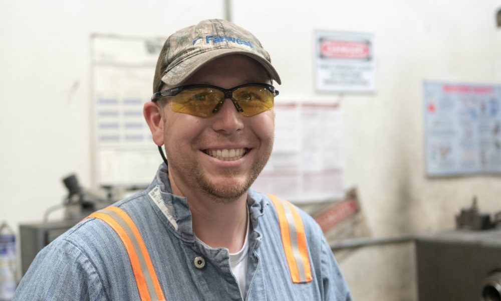 Worker smiling in industrial setting wearing safety glasses and a uniform with orange reflective str.