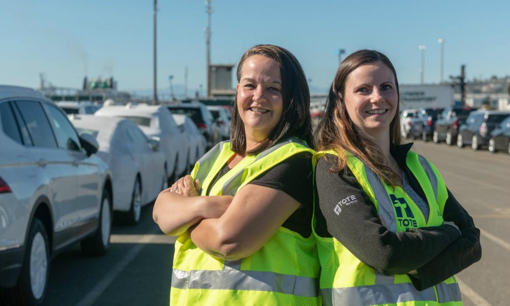 Two women in yellow safety vests standing back-to-back in a parking lot with cars and blue sky.