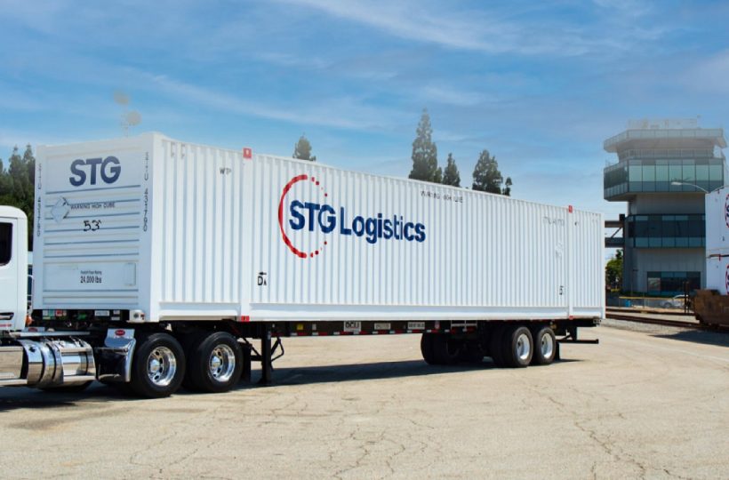 STG Logistics truck with containers on railway tracks under a blue sky.